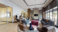 Located at 1900 Chapman Ave in Rockville, Maryland, this single-floor communal space is designed with a contemporary aesthetic that balances comfort and sophistication. The main lounge area, anchored by a prominent "GATHER" wall display, offers an open, airy layout with high ceilings and floor-to-ceiling windows that flood the space with natural light and provide views of the surrounding streetscape. The lounge is furnished with an eclectic mix of seating options, including a tufted leather daybed, plush sofas, accent chairs, and velvet ottomans, all arranged to encourage both socializing and quiet relaxation. Warm wood-paneled columns with open shelving display curated décor accents, while a wall-mounted flat-screen TV and a sculptural pendant chandelier add functional and visual interest. Light-toned herringbone flooring runs throughout, complementing the neutral and earthy color palette. A private dining room adjoins the lounge, featuring a large dark wood conference-style table surrounded by upholstered olive-toned chairs, set atop a bold patterned tile floor. A warm wood ceiling, a striking multi-arm chandelier, decorative convex mirror wall art, and a built-in media wall complete the refined yet inviting atmosphere of this dining area.