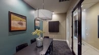 This single-story conference room offers a polished, professional environment designed for focused gatherings and meetings. The space is defined by a striking two-tone color scheme, pairing warm beige walls with bold teal accent walls that lend the room a contemporary, sophisticated character. A large, dark-finished conference table serves as the centerpiece, surrounded by matching black ergonomic chairs that comfortably accommodate multiple attendees.

Overhead, a prominent drum-style pendant light with a chrome frame provides warm, even illumination, complemented by recessed ceiling lights that enhance visibility throughout the room. The flooring features a richly patterned carpet in deep blues and earth tones, adding texture and visual interest underfoot.

Along one wall, a sleek dark credenza holds decorative accents including tall sculptural vases and a smooth stone piece, while a mounted dark-wood cabinet on the opposing wall offers additional storage. A glass-paneled door along one side allows access and contributes a sense of openness. Multiple framed abstract artworks in dark wood frames are displayed across the teal walls, reinforcing the room's curated aesthetic. The overall ambiance is refined, professional, and thoughtfully designed to support a productive meeting environment.