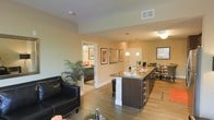 This single-floor residence offers a thoughtfully designed open-concept layout that integrates the living, dining, and kitchen areas into one cohesive and functional space. The living area is anchored by sleek hardwood-style flooring that extends throughout, creating a warm and unified aesthetic. A comfortable seating arrangement centers the living space, complemented by decorative framed artwork adorning the walls and a lush indoor plant that adds a natural touch.

The kitchen is a standout feature, boasting a large island with dark cabinetry and granite countertops, providing ample prep and entertaining space. Bar-height seating lines the island, while elegant pendant lighting above adds a refined ambiance. The kitchen flows naturally into a dedicated dining area, ideal for gatherings.

The warm, neutral wall tones throughout contribute to a welcoming atmosphere, while the open sightlines between rooms enhance the sense of spaciousness. A hallway visible from the main living area leads to additional rooms, suggesting a well-organized and private bedroom wing. Ceiling vents and recessed lighting ensure year-round comfort and consistent illumination. Overall, this space combines modern finishes with a practical, open layout to deliver a comfortable and stylish living environment.