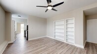This single-floor unit presents a clean, neutral aesthetic throughout, with light wood-style laminate flooring and warm greige walls creating a cohesive, modern feel across all rooms. Ceiling fans with integrated lighting are installed in multiple rooms, providing both air circulation and ambient light.

The main living area is a generously sized, open room featuring two sets of built-in white shelving units recessed into the wall — a practical and visually appealing storage solution. The space opens toward the kitchen, which is outfitted with white shaker-style cabinetry, black hardware, stone-look countertops, a stainless steel sink, a gas range, and a classic white subway tile backsplash that adds a timeless character.

The bedrooms are well-proportioned, with one featuring a standard closet with bifold doors and another offering window-unit climate control alongside a wall-mounted heating unit. A door from one bedroom provides direct access to an outdoor balcony. The bathroom is finished with white penny tile flooring, a full tub-shower combination, a white vanity, and a framed mirror with warm sconce lighting, completing the unit's clean and functional design.