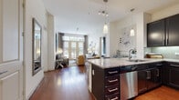 This well-appointed single-floor residence offers a thoughtfully designed open-concept layout that seamlessly connects the kitchen, dining, and living areas. Rich hardwood flooring flows throughout, anchoring the space with warmth and continuity.

The kitchen is a standout feature, outfitted with dark espresso cabinetry, granite countertops, a glass tile backsplash, and a full suite of stainless steel appliances including a gas range, microwave, refrigerator, and dishwasher. A central island with pendant lighting provides additional prep space and casual seating.

The living room is bright and inviting, benefiting from soaring ceilings and a wall of French doors that flood the space with natural light and open to an outdoor balcony with urban views. The room offers generous proportions ideal for relaxed entertaining.

The primary bedroom features rich hardwood floors, a dramatic upholstered headboard, track lighting, a large window with outdoor views, and access to a walk-in closet with built-in shelving. A second bedroom with a distinctive textured accent wall and natural light completes the sleeping quarters. Both bedrooms are served by well-finished bathrooms featuring dark cabinetry, granite vanities, and tile flooring.

A welcoming entry hallway with a large decorative mirror leads smoothly into the main living areas, completing this cohesive and elegantly appointed residence.