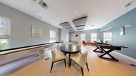 This single-floor amenity space functions as a well-equipped social and recreation lounge with a clean, modern aesthetic. The open layout is finished with light hardwood flooring and neutral gray walls, punctuated by a bold teal accent wall that adds visual interest near the entrance. Recessed lighting and pendant fixtures illuminate the space throughout, complemented by abundant natural light streaming through multiple windows fitted with roller shades.

The recreation area is the focal point, housing a contemporary white-framed pool table with overhead linear pendant lighting, a sleek stainless shuffleboard table, and a foosball table. A round dark-topped table with upholstered cream chairs provides a casual gathering spot centrally located within the room. A wall-mounted flat-screen television is positioned for easy viewing from the seating area.

A dedicated "Social Hub" workstation nook is outfitted with a desktop computer and printer, offering a functional workspace alongside the leisure amenities. A vibrant red accent chair adds a pop of color near the sun-filled windows of this nook. The space is enclosed by glass sliding doors, providing both visual openness and separation from adjacent areas. The overall ambiance is bright, inviting, and designed to encourage social interaction and leisure activities.