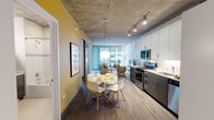 Located in Chicago's vibrant Loop neighborhood at 56 E Randolph St, this single-floor unit showcases a contemporary urban design defined by striking exposed concrete ceilings throughout. The open-concept living, dining, and kitchen area is anchored by floor-to-ceiling windows that flood the space with natural light and offer views of the surrounding cityscape.

The kitchen is well-appointed with stainless steel appliances, including a gas range, microwave, dishwasher, and refrigerator. Two-tone cabinetry pairs white upper cabinets with charcoal lower units, complemented by light countertops and a subway tile backsplash. A stacked in-unit washer and dryer is conveniently tucked away nearby.

The dining area accommodates a round table with seating for four, while the living room provides comfortable space for lounging with direct access to natural light from the expansive window wall. Wood-style flooring runs throughout the main living areas, adding warmth against the industrial ceiling.

The bedroom features exposed concrete ceilings, dark hardwood floors, and a spacious walk-in closet with built-in shelving and a full-length hanging rod. The bathroom includes a dark-vanity with a mirrored medicine cabinet and a tiled soaking tub and shower combination.