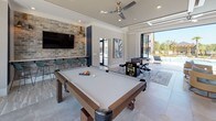 Located at 205 River Place in Augusta, Georgia, this single-story community clubhouse is thoughtfully designed to deliver an upscale recreational experience. The space is defined by its contemporary aesthetic, featuring light-toned tile flooring, coffered and tray ceilings with recessed lighting, and ceiling fans throughout.

The game room serves as a central highlight, equipped with a full-size billiards table, a foosball table, and an arcade game cabinet, all set against a striking exposed brick accent wall with a wall-mounted flat-screen television. Large floor-to-ceiling windows and expansive glass panel doors flood the space with natural light while providing direct views of the surrounding property.

The lounge area offers comfortable wicker seating with teal cushions arranged around low tables, anchored by a patterned area rug and accompanied by a wall-mounted television. Floor-to-ceiling bi-fold glass doors open entirely to the outdoor pool terrace, creating a seamless indoor-outdoor flow ideal for social gatherings.

The kitchen features a generous marble-topped island with bar seating, dark cabinetry, open white shelving with a textured stone backsplash, stainless steel appliances, and dramatic pendant lighting. A skylight above enhances the bright, welcoming atmosphere. Together, these spaces form a cohesive, amenity-rich environment designed for community engagement and leisure.