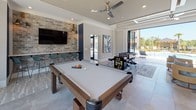 Located at 205 River Place in Augusta, Georgia, this single-story community clubhouse is thoughtfully designed to deliver an upscale recreational experience. The space is defined by its contemporary aesthetic, featuring light-toned tile flooring, coffered and tray ceilings with recessed lighting, and ceiling fans throughout.

The game room serves as a central highlight, equipped with a full-size billiards table, a foosball table, and an arcade game cabinet, all set against a striking exposed brick accent wall with a wall-mounted flat-screen television. Large floor-to-ceiling windows and expansive glass panel doors flood the space with natural light while providing direct views of the surrounding property.

The lounge area offers comfortable wicker seating with teal cushions arranged around low tables, anchored by a patterned area rug and accompanied by a wall-mounted television. Floor-to-ceiling bi-fold glass doors open entirely to the outdoor pool terrace, creating a seamless indoor-outdoor flow ideal for social gatherings.

The kitchen features a generous marble-topped island with bar seating, dark cabinetry, open white shelving with a textured stone backsplash, stainless steel appliances, and dramatic pendant lighting. A skylight above enhances the bright, welcoming atmosphere. Together, these spaces form a cohesive, amenity-rich environment designed for community engagement and leisure.