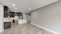 This single-floor, 2-bedroom, 1-bathroom residence offers a well-designed open-concept layout with a clean, modern aesthetic throughout. Light wood-look flooring and neutral gray walls create a cohesive, contemporary feel across all living areas.

The open living area is spacious and bright, featuring recessed lighting, a bold black-framed entry door with glass panels, and a large sliding glass door that allows abundant natural light and outdoor views. The kitchen is a standout feature, showcasing floor-to-ceiling dark wood-grain cabinetry with sleek hardware, white quartz countertops, a central island, tile backsplash, and stainless steel appliances — all illuminated by recessed ceiling lights.

The primary bedroom is generously sized, equipped with a ceiling fan and natural light from a window, and includes access to a dedicated walk-in closet with multi-tiered wire shelving. The second bedroom also features a ceiling fan and a window with outdoor views. Two entrance points provide added convenience and accessibility.

The bathroom is finished with marble-style tile, a white quartz vanity with an illuminated mirror, and a soaking tub with tiled surround. A dedicated utility room and additional closet complete the functional layout of this well-appointed residence.