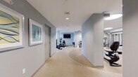 Located at 2432 Albany Ave in West Hartford, CT, this fitness center offers a comprehensive, well-organized workout facility across a single floor. The space is designed with a clean, modern aesthetic, featuring light gray walls, neutral speckled flooring, and bright recessed and linear ceiling lighting that creates an open, energizing atmosphere.

The facility is divided into distinct functional zones that flow seamlessly from one to the next. The main cardio area houses a variety of Life Fitness machines, including ellipticals, upright and recumbent bikes, and a stair climber, all arranged to allow comfortable spacing between equipment. Large circular ceiling fixtures illuminate this section, which also features wall-mounted mirrors that enhance the sense of openness.

A dedicated strength training zone is equipped with a multi-station cable machine, a full dumbbell rack with accompanying benches, and additional resistance equipment, all anchored by a wall-mounted flat-screen TV. Adjacent to this is a cycling studio featuring two smart stationary bikes with monitor screens, set on a curved floor design, along with shelving stocked with foam rollers and fitness accessories for flexibility and recovery work.

A corridor connects the zones, accented with framed artwork that adds a welcoming touch. The thoughtful layout, modern equipment selection, and clean finishes make this a functional and inviting fitness destination.