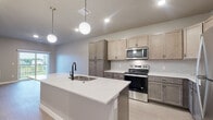 This single-floor space is designed with a contemporary aesthetic and a functional open-concept layout. The kitchen serves as the heart of the unit, showcasing two-tone cabinetry in warm taupe and darker lower tones, complemented by a large quartz-topped island with a matte black sink faucet. Stainless steel appliances, including a range, microwave, and refrigerator, are paired with a neutral tile backsplash. Globe pendant lights hang above the island, adding a modern touch alongside recessed ceiling lighting.

The living area connects seamlessly to the kitchen and features light wood-look flooring, neutral gray walls, and a sliding glass door that opens to a private balcony with outdoor views. A wall-mounted TV bracket is pre-installed for convenience.

The bedrooms are finished with plush carpeting and feature windows that bring in natural light and outdoor views. A walk-in closet with wire shelving offers ample storage and connects directly to a bathroom. The bathroom includes a tub-shower combination with matte black fixtures. A dedicated laundry area with a full-size washer and dryer adds practical convenience to the space.