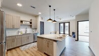 This contemporary single-floor residence offers a thoughtfully designed open-concept layout that seamlessly connects the kitchen, dining, and living areas. The kitchen serves as the centerpiece of the space, showcasing light wood cabinetry, a white subway tile backsplash, and a granite-topped island illuminated by a pair of stylish pendant lights. Stainless steel appliances — including a refrigerator, microwave, and dishwasher — complement the modern aesthetic, while light wood-tone vinyl plank flooring runs throughout the main living areas, adding warmth and cohesion. A ceiling fan anchors the living area, which is bathed in natural light from large French doors that open to an outdoor space. Stylish paneled interior doors in a charcoal-gray finish provide a sophisticated contrast to the neutral walls throughout. The bathroom features granite countertops, light wood cabinetry, and a tub-shower combination with marble-style tile surround, all set on matching wood-tone flooring. The bedrooms offer plush carpeting underfoot, generous wall space, and windows that invite natural light. A walk-in closet adds practical storage. The overall ambiance is clean, bright, and modern, combining functional design with contemporary finishes throughout.