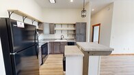 Located in Bloomington, Indiana, this thoughtfully designed single-floor unit offers three bedrooms and three full bathrooms within a well-utilized footprint. The kitchen serves as a standout centerpiece, showcasing gray-toned cabinetry, open wood shelving with black iron brackets, light stone countertops, black appliances, and a central island with a white base — all set atop warm light hardwood flooring. A pendant lantern-style light fixture adds a rustic-modern touch, with high ceilings amplifying the sense of openness. The bedrooms feature plush carpeting and warm wood baseboards and trim. The primary bedroom is highlighted by elegant arched windows that flood the room with natural light and offer views of the surrounding greenery. Additional bedrooms include ceiling fans and windows that continue the bright, airy feel. Each of the three bathrooms is finished with earthy stone tile flooring, dark-framed mirrors, bronze fixtures, and rich wood vanities topped with white countertops, creating a cohesive and refined aesthetic. Arched windows appear throughout, lending architectural character to the space. The overall ambiance is warm and contemporary, blending practical design with inviting details throughout.