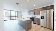 This contemporary one-bedroom, one-bathroom unit offers a well-designed open-concept layout across a single floor. The combined kitchen and living area serves as the heart of the space, anchored by a generous kitchen island with a white quartz countertop and an integrated sink. Rich dark-toned cabinetry lines the walls alongside a classic white subway tile backsplash, complemented by a full suite of stainless steel appliances including a refrigerator with ice dispenser, electric range, and microwave. Recessed lighting and large-format tile flooring throughout lend a clean, modern aesthetic. The living area opens to expansive floor-to-ceiling windows that frame striking panoramic city views, flooding the space with natural light. The spacious bedroom continues the floor-to-ceiling window theme, featuring an exposed concrete structural column that adds an industrial character, along with views of the surrounding urban skyline. A dedicated walk-in closet offers built-in shelving and hanging rods for ample storage. The bathroom is finished with white subway tile in a glass-enclosed walk-in shower, complemented by warm-toned tile flooring. Throughout the unit, the consistent use of neutral tones, large-format tile, and modern finishes creates a cohesive and refined living environment.
