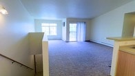 This single-floor space offers a functional and straightforward layout distributed across several interconnected rooms. The 3D floor plan reveals a well-organized arrangement that includes a spacious living area, a kitchen, and multiple additional rooms connected by a central corridor.

The living area is notably generous in scale, featuring wall-to-wall carpeting in a neutral tone that extends throughout the main living spaces. Two windows allow ample natural light to fill the room, complemented by a sliding glass door that opens to an outdoor balcony, enhancing the sense of openness and providing outdoor access. Baseboard heating units run along the exterior walls, offering an efficient heating solution throughout the space.

The kitchen is positioned toward the interior of the unit and is separated from the living area by a partial wall, creating a defined yet connected flow between spaces. Additional rooms branch off from the main hallway, each with standard doorways and neutral wall finishes. The overall aesthetic is clean and minimal, with white walls throughout that contribute to a bright, open ambiance well-suited for personalization.