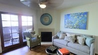 This single-floor space offers a welcoming and thoughtfully curated living environment that blends comfort with artistic personality. The living room serves as the heart of the space, anchored by a classic fireplace with a clean, neutral surround that adds warmth and a timeless focal point to the room. Wood flooring runs throughout, lending a warm, natural tone that complements the overall aesthetic.

The seating area is well-appointed with a plush gray sofa dressed in an assortment of patterned and solid accent pillows, paired with a charming green armchair adorned with a cozy faux-fur throw. A round coffee table styled with books and decorative objects sits at the center, adding a lived-in, inviting quality.

The walls are adorned with standout artwork, including a large, vibrant painting of blossoming branches in shades of blue and white, and a striking circular mosaic wall piece that adds a bohemian touch. A textured popcorn ceiling features an ornate ceiling medallion framing a central light fixture, adding a subtle classical detail overhead.

Natural light filters in through a wood-framed window and an adjacent door, hinting at outdoor access. The overall ambiance is eclectic, cozy, and rich with personal expression, making this a comfortable and visually engaging living space.