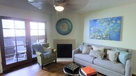 This single-floor space offers a welcoming and thoughtfully curated living environment that blends comfort with artistic personality. The living room serves as the heart of the space, anchored by a classic fireplace with a clean, neutral surround that adds warmth and a timeless focal point to the room. Wood flooring runs throughout, lending a warm, natural tone that complements the overall aesthetic.

The seating area is well-appointed with a plush gray sofa dressed in an assortment of patterned and solid accent pillows, paired with a charming green armchair adorned with a cozy faux-fur throw. A round coffee table styled with books and decorative objects sits at the center, adding a lived-in, inviting quality.

The walls are adorned with standout artwork, including a large, vibrant painting of blossoming branches in shades of blue and white, and a striking circular mosaic wall piece that adds a bohemian touch. A textured popcorn ceiling features an ornate ceiling medallion framing a central light fixture, adding a subtle classical detail overhead.

Natural light filters in through a wood-framed window and an adjacent door, hinting at outdoor access. The overall ambiance is eclectic, cozy, and rich with personal expression, making this a comfortable and visually engaging living space.