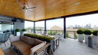 This rooftop amenity space seamlessly blends indoor comfort with open-air living through a thoughtfully designed layout that transitions between enclosed and covered outdoor areas. Throughout, warm tongue-and-groove wood ceilings lend a cohesive, inviting character, while large-format porcelain tile flooring unifies the space underfoot.

The indoor lounge area is lined with expansive windows fitted with roller shades, allowing abundant natural light while offering privacy control. Dark leather seating arrangements, including sofas and swivel accent chairs centered around glass-top tables, create a relaxed yet polished atmosphere. Lush potted plants, including a prominent fiddle-leaf fig, add organic warmth to the interior.

A built-in bar counter runs along a folding window wall that opens directly to panoramic city skyline views, accompanied by teal metal barstools — an ideal perch for casual gatherings. Adjacent, an indoor lounge area features a mounted flat-screen television above a linear fireplace surround, flanked by painted brick walls and contemporary seating.

The covered outdoor terrace extends the living space further, featuring ceiling fans overhead and sleek cable-rail balcony fencing that preserves sweeping urban views. Neutral upholstered outdoor furniture, natural wood stump accent tables, and lush planters complete the al fresco setting, offering a sophisticated environment for lounging and entertaining.
