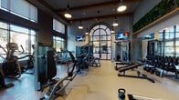 This modern fitness center is thoughtfully designed to support a full range of workout routines across multiple distinct zones. The main strength and cardio area boasts soaring ceilings with exposed wood beam accents, pendant lighting, and ceiling fans, creating an airy yet energetic atmosphere. A dramatic arched steel-frame window serves as a striking architectural focal point, flooding the space with natural light while offering views of the outdoor courtyard. Multiple wall-mounted TVs are positioned throughout for convenience during workouts. The strength zone is equipped with a comprehensive selection of cable machines, free weights, benches, and kettlebells, while a row of treadmills lines one wall. A large mirrored wall amplifies the sense of space and supports form-checking during exercise. Adjacent to the main floor, a glass-walled cycling and group fitness studio features herringbone wood-look flooring, stationary bikes, interactive fitness screens, and a full mirrored wall with a large display screen. A lush living green wall adorns the upper interior, adding a vibrant natural element. A dedicated amenity nook near the entrance offers yoga mats, storage baskets, a beverage cooler, and counter space, ensuring members have everything needed for a seamless workout experience. Floor-to-ceiling windows throughout provide views of the surrounding greenery.