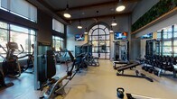 This modern fitness center is thoughtfully designed to support a full range of workout routines across multiple distinct zones. The main strength and cardio area boasts soaring ceilings with exposed wood beam accents, pendant lighting, and ceiling fans, creating an airy yet energetic atmosphere. A dramatic arched steel-frame window serves as a striking architectural focal point, flooding the space with natural light while offering views of the outdoor courtyard. Multiple wall-mounted TVs are positioned throughout for convenience during workouts. The strength zone is equipped with a comprehensive selection of cable machines, free weights, benches, and kettlebells, while a row of treadmills lines one wall. A large mirrored wall amplifies the sense of space and supports form-checking during exercise. Adjacent to the main floor, a glass-walled cycling and group fitness studio features herringbone wood-look flooring, stationary bikes, interactive fitness screens, and a full mirrored wall with a large display screen. A lush living green wall adorns the upper interior, adding a vibrant natural element. A dedicated amenity nook near the entrance offers yoga mats, storage baskets, a beverage cooler, and counter space, ensuring members have everything needed for a seamless workout experience. Floor-to-ceiling windows throughout provide views of the surrounding greenery.