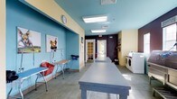 This single-floor facility is designed to accommodate pet grooming and care services in a clean, well-organized environment. The space is immediately distinguished by its vibrant color palette, combining sky-blue ceilings with soft yellow walls and a deep plum accent wall toward the rear, creating a welcoming yet professional atmosphere.

The main area centers around a large grooming table with a durable gray surface, positioned prominently for easy access. A secondary grooming station along the side wall is equipped with a grooming arm, blow dryer, and storage basket, providing a fully functional workstation. Tile flooring throughout ensures easy maintenance and hygiene.

Recessed fluorescent panel lighting keeps the space evenly and brightly illuminated, complementing the natural light that filters in through the glass-paneled door at the far end of the corridor. The hallway leads toward what appears to be an exterior entrance, adding convenient accessibility.

To one side, a dedicated laundry area houses a washer and dryer, supporting the facility's operational needs. Decorative pet-themed artwork on the walls adds personality, while a wall clock and organized supplies reflect a practical, detail-oriented setup throughout.