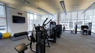 This well-appointed fitness center offers a bright, open workout environment designed to support a full range of exercise needs. The space is anchored by expansive floor-to-ceiling windows that line the perimeter, flooding the interior with abundant natural light and providing sweeping views of the surrounding urban skyline — an energizing backdrop for any workout.

The facility is outfitted with a comprehensive selection of professional-grade strength training machines, including multi-function cable and resistance units positioned throughout the floor. Cardio equipment is also present, complementing the strength-focused stations. The layout is thoughtfully organized to allow comfortable circulation between machines.

Overhead, sleek linear pendant lighting supplements the natural light, ensuring consistent illumination throughout the space. The neutral-toned carpet tiles contribute to a clean, polished aesthetic while providing comfort underfoot. A wall-mounted flat-screen television and a digital clock are positioned for easy visibility, adding functional amenities to the workout experience.

The overall ambiance is modern and professional, with white walls, structural columns, and clean architectural lines reinforcing the contemporary design. This fitness center is well-suited for users seeking a bright, fully equipped space with an inspiring urban outlook.