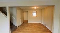 This two-story home offers a clean, functional layout with thoughtful design details throughout. The main level is anchored by warm hardwood flooring that extends seamlessly from the living area through to the entryway, lending a sense of continuity and warmth to the space. Neutral beige walls provide a versatile backdrop, complementing the natural tones of the flooring and creating a bright, welcoming atmosphere.

A staircase with carpeted treads is conveniently positioned along one side of the main level, providing access to the upper floor while leaving the living space open and unobstructed. A flush-mount ceiling light fixture provides overhead illumination, ensuring the interior remains well-lit. A small window cutout in the wall introduces natural light into the space, adding an architectural detail that enhances the sense of openness.

The entryway, visible from the living area, features a paneled front door flanked by a closet, offering practical storage right at the entrance. The overall layout is straightforward and efficient, with clean lines and minimal ornamentation that make the space easy to personalize and furnish to suit a variety of needs.