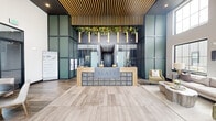 The Slate lobby is a thoughtfully designed single-floor space that makes a strong architectural statement upon entry. The centerpiece is a striking reception desk finished with a textured tile face and warm wood surround, prominently branded with the Slate name. Overhead, a dramatic slatted wood ceiling treatment runs the length of the space, complemented by cylindrical pendant lights and cascading greenery that introduce a natural, organic element against the dark ceiling backdrop. Flanking the reception area, floor-to-ceiling glass panel walls with a grid pattern in muted sage green create visual depth and define zones within the open layout. The entry foyer features soaring double-height ceilings flooded with natural light from large multi-pane windows, offering views to the exterior. A comfortable lounge area includes a neutral sofa, olive green accent chairs, and an organic-shaped stone coffee table arranged on a soft area rug. A secondary seating nook with a round table, upholstered dining chairs, and a wall-mounted display provides a functional gathering area. Light wood-look tile flooring flows consistently throughout, unifying the space with a clean, contemporary aesthetic.