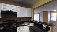 This single-floor residential unit offers a practical and well-organized layout designed for comfortable everyday living. The kitchen is a standout feature, showcasing dark granite-style countertops that contrast sharply with crisp white raised-panel cabinetry above and below. A stainless steel double-basin sink is set into the corner of the countertop, providing ample workspace on either side. Black appliances, including an electric range and dishwasher, complete the kitchen's cohesive color scheme. A textured ceiling adds a classic touch overhead. One of the kitchen's most notable architectural features is the pass-through window that opens directly into the adjacent living area, creating a sense of openness and connectivity between the two spaces. Warm golden-toned walls run throughout, lending a cozy and inviting ambiance to the interior. The living area, visible through the pass-through, is brightened by a large window dressed with vertical blinds, allowing natural light to filter into the space. The overall design balances function and comfort, making efficient use of the available space while maintaining a clean, classic aesthetic throughout the home.