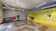 This single-floor fitness studio is thoughtfully designed to support a wide range of workout activities. The space is immediately distinguished by its striking hexagonal-patterned flooring in neutral tones of tan and grey, providing both visual interest and a durable, functional surface suited for high-intensity use. One side of the studio features a raw, exposed concrete wall that lends an industrial character, contrasting effectively with the bold yellow-green accent walls on the opposite end, which are lined with full-length mirrors to enhance the sense of space and support form-focused training.

Overhead, exposed ductwork and piping run along the white ceiling, complemented by linear LED pendant lighting that ensures bright, even illumination throughout. Large frosted windows along one wall allow natural light to filter into the space while maintaining privacy. The studio is well-equipped with kettlebells, a dumbbell rack, stability balls on a dedicated rack, medicine balls, a BOSU balance trainer, an adjustable weight bench, and a power tower for bodyweight exercises. The open floor plan allows for flexible arrangement, making the space suitable for group fitness classes, personal training, or independent workouts.