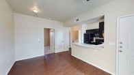 This single-floor unit showcases an open-concept design that integrates the kitchen and living area into a cohesive, functional space. The flooring throughout the main living area is rich, dark hardwood, adding warmth and visual depth to the neutral, cream-toned walls.

The kitchen is positioned along one side of the open layout, featuring a raised breakfast bar peninsula that provides a natural division between cooking and living spaces. Black appliances, including a refrigerator, range, and over-the-range microwave, are paired with dark cabinetry, creating a bold contrast against the light countertops and walls. A sink is conveniently integrated into the peninsula countertop.

The living area is generously proportioned, with a ceiling fan overhead and ample wall space. Doorways visible from the main area lead to additional rooms, including what appears to be a carpeted bedroom and a laundry area with a washer and dryer, suggesting strong functional utility throughout. HVAC vents are consistently placed for efficient climate control. The overall ambiance is clean, practical, and move-in ready, with a straightforward layout designed for everyday comfort.