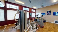 This fitness facility offers a comprehensive workout environment designed to support a wide range of exercise routines. The main gym area features warm hardwood-style flooring and a bold two-tone color scheme, with deep crimson accent walls contrasting against neutral beige tones. High clerestory windows line the upper portion of the walls, allowing natural light to filter in while maintaining privacy, and ceiling fans help circulate air throughout the space.

The gym is outfitted with a diverse selection of equipment, including multi-station strength machines, cable weight systems, treadmills, elliptical trainers, stair climbers, and stationary bikes. Full-length mirrors along one wall visually expand the space and allow users to monitor their form during workouts. A wall-mounted television provides entertainment, and motivational sports and nature photography decorates the walls, adding energy to the environment.

Adjacent to the fitness room is a serene indoor swimming pool enclosure. This space features a vaulted ceiling with a row of skylights that flood the area with natural light, creating a bright and airy atmosphere. Elegant arched windows with decorative leaded glass line the walls, and lush hanging plants add a touch of greenery. Lounge chairs are arranged along the pool deck on both sides, offering a comfortable space to relax before or after a swim.