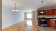 This single-floor residence on Crown Court offers a well-designed, open-concept layout that promotes a sense of spaciousness and easy flow between living areas. The main living and dining space is anchored by warm, polished hardwood flooring that reflects natural light streaming in from a large window at the far end of the room. A classic multi-arm chandelier adds a touch of elegance to the dining area, complementing the neutral, light-toned walls that create a bright and welcoming ambiance throughout. The kitchen is positioned in an open, accessible arrangement adjacent to the living area, featuring rich warm-toned cabinetry paired with dark countertops for a pleasing contrast. Stainless steel appliances, including a gas range and an over-the-range microwave, provide a modern, functional edge. The overall design balances practicality with comfort, making the space well-suited for everyday living and entertaining alike. Clean white baseboards, neutral wall tones, and consistent flooring throughout contribute to a cohesive and polished aesthetic across the entire floor.