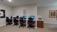 Located at 201 Kroger Drive in Richmond Hill, Georgia, this single-floor community facility offers a thoughtfully designed layout with a variety of functional spaces to serve its residents. The heart of the facility is a generous open-concept common area, where a comfortable lounge with plush blue seating, a mounted flat-screen TV, and ceiling fans creates a welcoming gathering space. Tray ceilings with crown molding and recessed lighting lend an elevated architectural character throughout. Adjacent to the lounge, multiple dining areas feature dark wood tables and upholstered chairs, ideal for shared meals and social gatherings. A dedicated fitness room is well-lit by large windows overlooking greenery and is equipped with treadmills, an elliptical machine, and wall mirrors. The computer center provides several individual workstations with desktop computers and a shared printer, supporting residents' connectivity needs. A private office area with built-in cabinetry and ample counter space offers a quiet workspace. Neutral wall tones, plush carpeting, and consistent décor throughout the facility create a cohesive, comfortable ambiance. Glass-paned entry doors and open transitional spaces ensure easy navigation across this single-level community hub.