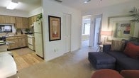This single-floor residence offers a practical and comfortable layout designed for easy everyday living. The living area is finished with neutral-toned carpet that extends throughout the main space, creating a warm and cohesive feel. A dark upholstered sofa and side lamp anchor the sitting area, which flows naturally toward a hallway leading to a well-lit bedroom, visible through an open doorway with light filtering in from a window.

Adjacent to the living space, the kitchen features light wood cabinetry, a white refrigerator, and a stove, providing a functional cooking environment. The transition between the kitchen's hard flooring and the carpeted living area adds a subtle visual distinction between the two spaces. A louvered closet door along the main wall offers convenient storage access, while a framed piece of wall art adds a decorative touch to the otherwise clean, understated interior.

The layout is straightforward and open, allowing for natural movement between areas. Neutral wall tones and simple finishes throughout give the space a clean and move-in-ready appearance, making it an accessible and functional residential unit suited for comfortable day-to-day living.