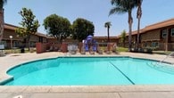 This single-story residential community is designed with comfort and outdoor living in mind. At the heart of the property lies a generously sized outdoor swimming pool with crystal-clear turquoise water, ideal for warm-weather relaxation. The pool deck is lined with lounge chairs, offering ample space for sunbathing and leisure. A brick partition wall adds a classic architectural touch, neatly framing the pool area and providing a sense of structure within the outdoor space.

Adjacent to the pool is a vibrant children's playground equipped with slides and climbing structures, making the community especially well-suited for families. The surrounding grounds are beautifully landscaped with mature palm trees and lush shade trees that provide natural canopy and a sense of privacy.

The residential buildings follow a consistent single-story design with an earth-toned exterior, blending seamlessly with the outdoor environment. The layout of the community encourages an open, accessible feel, with well-maintained walkways and green lawn areas connecting the various amenities. The overall ambiance is one of a relaxed, sun-drenched community that prioritizes outdoor activity, social gathering, and everyday convenience in a welcoming and well-kept setting.