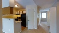 This single-floor residential unit offers a practical and comfortable layout designed for everyday living. The kitchen is positioned at the heart of the home, featuring warm wood-toned upper and lower cabinetry with white panel insets, a black refrigerator, electric range, built-in microwave, and a raised breakfast bar that provides a natural division between the kitchen and the adjoining living areas. The flooring in the kitchen is hard surface, transitioning seamlessly to plush wall-to-wall carpeting throughout the hallway and living spaces beyond. A central hallway extends from the kitchen area, lined with doors that provide access to additional rooms, and terminates at a bright window that draws natural light deep into the interior. The neutral color palette of white walls and light grey carpet creates a clean, airy atmosphere throughout. Ceiling-mounted light fixtures provide consistent ambient lighting, and a fire sprinkler system is visible, reflecting attention to safety. The overall layout is efficient and functional, with clearly defined zones for cooking, dining, and living, making the space well-suited for comfortable day-to-day use.
