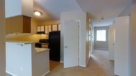 This single-floor residential unit offers a practical and comfortable layout designed for everyday living. The kitchen is positioned at the heart of the home, featuring warm wood-toned upper and lower cabinetry with white panel insets, a black refrigerator, electric range, built-in microwave, and a raised breakfast bar that provides a natural division between the kitchen and the adjoining living areas. The flooring in the kitchen is hard surface, transitioning seamlessly to plush wall-to-wall carpeting throughout the hallway and living spaces beyond. A central hallway extends from the kitchen area, lined with doors that provide access to additional rooms, and terminates at a bright window that draws natural light deep into the interior. The neutral color palette of white walls and light grey carpet creates a clean, airy atmosphere throughout. Ceiling-mounted light fixtures provide consistent ambient lighting, and a fire sprinkler system is visible, reflecting attention to safety. The overall layout is efficient and functional, with clearly defined zones for cooking, dining, and living, making the space well-suited for comfortable day-to-day use.