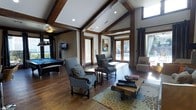 This single-floor space makes an immediate impression with its dramatic vaulted ceilings adorned with rich, dark-stained exposed wood beams that run the length of the room, adding both architectural character and a warm, lodge-inspired aesthetic. Hardwood flooring flows throughout, complementing the wood-toned trim and structural accents that frame the doorways and walls.

The main lounge area is thoughtfully arranged with classic upholstered armchairs set around a layered wood coffee table atop a vibrant floral-patterned area rug, creating an inviting and comfortable gathering space. Natural light pours in generously through large French doors and tall windows, offering views of an outdoor pool area and flooding the interior with warmth.

A dedicated billiards zone anchors one side of the room, complete with a full-sized pool table and a wall-mounted television, making it an ideal entertainment hub. Recessed lighting installed within the vaulted ceiling supplements the abundant natural light, ensuring the space remains bright and welcoming at all hours.

The overall ambiance strikes a balance between casual comfort and refined design, with the combination of soaring ceilings, natural materials, and ample glazing lending the space an open, airy, and inviting character well-suited for socializing and recreation.