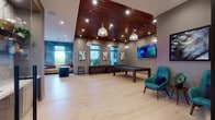 Located at 3991 NW 82nd Dr in Cooper City, Florida, this single-floor community clubhouse is thoughtfully designed to offer an engaging and upscale social environment. The space is anchored by a spacious game room showcasing rich light hardwood flooring, striking wood-paneled ceilings with recessed lighting, and pendant light fixtures that lend a warm, contemporary ambiance. The game area is equipped with a pool table, shuffleboard table, and a classic arcade pinball machine, complemented by a large wall-mounted flat-screen television — making it an ideal gathering spot for recreation and entertainment. Textured vertical-stripe wall panels and curated botanical artwork add refined detail throughout. A comfortable lounge area features a sofa, ottomans, and accent chairs in vibrant teal upholstery set against a dramatic teal-painted feature wall, with large windows that invite abundant natural light and views of the surrounding greenery. A dedicated wet bar area showcases dark wood cabinetry, crisp white quartz countertops, an eye-catching marble-look backsplash, open display shelving, and an under-counter beverage refrigerator — all accessible through a sleek glass-paneled door. Near the entrance, a welcoming seating nook with two teal accent chairs and a textured accent wall creates an inviting transition into the space.