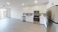 This single-floor unit offers a thoughtfully designed layout with a clean, contemporary aesthetic carried throughout every room. The open-concept kitchen is a standout feature, showcasing floor-to-ceiling white cabinetry, a textured gray tile backsplash, white quartz countertops, and a full suite of stainless steel appliances including a refrigerator, range, microwave, and dishwasher. Light-toned hardwood-style flooring runs through the kitchen and living areas, enhancing the bright and airy feel created by recessed lighting overhead.

The bedroom is carpeted for comfort and includes a ceiling fan with a window allowing natural light into the space. A generously sized walk-in closet serves the bedroom, equipped with adjustable wire shelving systems and multiple hanging rods across two walls, offering ample organization potential.

The bathroom is finished in a crisp, all-white palette with large-format tile flooring, an extended vanity featuring white cabinetry and a wide mirror, brushed nickel hardware, and a walk-in shower. The overall design of the space emphasizes modern minimalism, with neutral tones, clean lines, and quality finishes creating a cohesive and functional living environment.