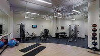 This single-floor fitness center is designed to support a comprehensive workout experience. The space features an open layout with high ceilings that expose industrial-style ductwork and piping, lending the gym a functional, utilitarian aesthetic. Bright fluorescent lighting runs throughout, ensuring the entire area is well-lit during any workout session.

The gym is outfitted with a solid selection of strength training equipment, including a multi-function cable machine, a leg press/curl machine, and an adjustable weight bench. A tiered dumbbell rack provides a range of free weight options to accommodate various fitness levels. A wall-mounted flat-screen television adds an entertainment element, making workouts more engaging.

Flooring transitions thoughtfully throughout the space — durable rubber matting covers the strength training zones, providing grip and impact absorption, while a smooth hardwood floor defines a dedicated stretching and yoga area at the front. Two exercise mats are laid out, ready for floor-based workouts or cool-down routines.

The neutral, cream-colored walls keep the space feeling clean and focused. Visible structural columns and exposed ceiling infrastructure contribute to the gym's industrial character. Overall, this fitness center offers a practical and well-organized environment suited for a full range of exercise routines.