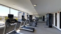 The fitness center at 100 Citronica in Lemon Grove is a well-appointed, single-level exercise facility designed to provide a functional and motivating workout environment. The space is lined with expansive floor-to-ceiling windows along one side, flooding the interior with abundant natural light while offering open views of the surrounding area, creating an airy and energizing atmosphere.

The facility is equipped with a comprehensive selection of cardio machines, including treadmills and elliptical trainers, arranged in an organized row that maximizes the available floor space. Full-length mirrored walls run along one side of the room, visually expanding the space and allowing users to monitor their form during exercise. The mirrors also reflect the natural light, further brightening the interior.

The ceiling features a clean, modern design with recessed and flush-mounted lighting to supplement natural light during all hours of use. The flooring is a durable, light-toned surface suited for high-traffic fitness use. Overall, the space presents a sleek, contemporary aesthetic that is both practical and welcoming for regular fitness activities.