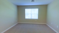 This single-floor space presents a clean and functional layout designed with simplicity and practicality in mind. The interior is finished with soft, neutral-toned walls in a warm beige palette that creates a calm and inviting atmosphere throughout. The room features wall-to-wall carpeting in a light, understated shade, providing a comfortable and quiet underfoot experience. A centered window fitted with white horizontal blinds allows natural light to filter into the room, offering a connection to the greenery visible outside while maintaining privacy. The white baseboard trim provides a crisp contrast against the neutral walls, adding a subtle architectural detail that gives the space a polished look. Ceiling-mounted HVAC venting is integrated discreetly into the ceiling, reflecting a thoughtfully designed mechanical layout. The overall ambiance of the space is quiet and unassuming, offering a versatile blank canvas that can be adapted to a variety of uses. The straightforward design and well-maintained finishes make this a functional and move-in ready environment suited for those seeking a comfortable and understated living or working space.