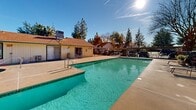Located at 849 Lincoln Rd in Yuba City, California, this outdoor amenity area serves as a central gathering space for the surrounding residential community. The centerpiece is a generously sized rectangular swimming pool with clear, turquoise water, complemented by a separate circular spa nestled nearby — ideal for relaxation year-round under California's sunny skies.

The pool deck is finished in smooth concrete, providing ample space for lounging and outdoor furniture, while a secure wrought iron fence encloses the entire perimeter, ensuring both safety and privacy. Entry points are accessible via poolside steps and handrails.

A single-story utility or clubhouse building sits adjacent to the pool area, finished with vertical wood siding in neutral tones and capped with a gabled roof — providing practical access to pool-related amenities. Covered carport parking is visible beyond the fencing, adding convenience for residents.

The surrounding landscape features a mature canopy of tall pine and cypress trees that frame the space, offering partial shade and a sense of natural seclusion. The open sky above allows for abundant natural light throughout the day, creating a bright and inviting outdoor environment well-suited for both leisure and community activity.
