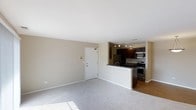 This single-floor space showcases an open-concept design that seamlessly connects the living area and kitchen. The living area is finished with soft, neutral-toned carpeting and painted in warm beige tones, creating a clean and inviting atmosphere. Natural light filters in, casting bright patches across the floor and enhancing the sense of openness.

The kitchen is partially open to the living space, divided by a raised breakfast bar that doubles as a casual dining or prep area. It is outfitted with stainless steel appliances, including a refrigerator, microwave, and range, all complemented by rich, dark cabinetry that provides a sharp contrast to the lighter surroundings. Track lighting installed on the kitchen ceiling offers focused illumination for the workspace. The kitchen floor transitions to hardwood-style flooring, distinguishing it from the carpeted living area.

A white paneled entry door is visible from the living room, providing a clear sense of the layout's flow. The overall design balances functionality with a modern aesthetic, making efficient use of the available space.
