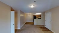 This single-floor unit offers an open-concept layout that seamlessly connects the living area, dining space, and kitchen. The living room is finished with wall-to-wall carpeting and neutral beige walls, creating a warm and understated atmosphere. A flush-mount ceiling light provides overhead illumination, while a smoke detector is also ceiling-mounted. White baseboards run throughout, adding a clean contrast to the flooring and walls.

The dining area transitions naturally from the living space and features a small chandelier, adding a touch of elegance. The flooring shifts to a hardwood-style surface in this zone, visually distinguishing it from the carpeted living area.

The kitchen is positioned at the rear of the open layout and is partially defined by a raised breakfast bar with a dark countertop. It is equipped with medium-toned wood cabinetry, a black refrigerator, and a range, all beneath dedicated overhead lighting. The textured popcorn ceiling is consistent throughout the unit. Two doors are visible — one at the front entry and one leading to an additional room — offering functional separation within the otherwise open floor plan.