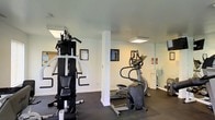 Located at 401 Bedford Lane in New Castle, Delaware, this community fitness center offers residents a dedicated space for exercise and wellness. The single-floor facility is designed with functionality in mind, featuring white walls, dark rubber flooring, and flush-mounted ceiling lighting that creates a bright and clean workout environment. The space is equipped with a multi-station strength training system and a cable weight machine, providing comprehensive resistance training options. Cardio equipment, including a stair climber and elliptical trainer, rounds out the exercise offerings. A structural column provides definition within the open layout, while a wall mirror enhances the sense of space and allows users to monitor their form during workouts. Safety is addressed with a visible fire extinguisher station. Framed artwork and informational postings on the walls, including a community notice board, add a welcoming touch to the utilitarian setting. The overall ambiance is practical and well-maintained, offering residents a convenient and functional fitness resource within their community.