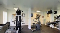 Located at 401 Bedford Lane in New Castle, Delaware, this community fitness center offers residents a dedicated space for exercise and wellness. The single-floor facility is designed with functionality in mind, featuring white walls, dark rubber flooring, and flush-mounted ceiling lighting that creates a bright and clean workout environment. The space is equipped with a multi-station strength training system and a cable weight machine, providing comprehensive resistance training options. Cardio equipment, including a stair climber and elliptical trainer, rounds out the exercise offerings. A structural column provides definition within the open layout, while a wall mirror enhances the sense of space and allows users to monitor their form during workouts. Safety is addressed with a visible fire extinguisher station. Framed artwork and informational postings on the walls, including a community notice board, add a welcoming touch to the utilitarian setting. The overall ambiance is practical and well-maintained, offering residents a convenient and functional fitness resource within their community.