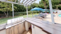 This outdoor space is thoughtfully designed for al fresco entertaining, centered around a fully equipped outdoor kitchen and a pristine swimming pool. The outdoor kitchen is the focal point, anchored beneath a clean-lined white pergola that provides partial shade while maintaining an open-air ambiance. The kitchen features a U-shaped stainless steel configuration with multiple built-in grills, generous counter space with smooth composite surfaces, and integrated cabinetry — offering everything needed for outdoor cooking and food preparation.

Surrounding the kitchen, the space opens onto a spacious patio deck that connects seamlessly to the pool area. The pool is generously sized and features a classic tiled border, with lounge chairs and vibrant blue umbrellas arranged poolside for comfortable relaxation. A balcony overlooks the pool from an adjacent structure, adding a sense of vertical dimension to the space.

The landscaping throughout is lush and well-maintained, with manicured lawns, neatly trimmed hedgerows, mature trees, and tropical palms that create a private, resort-like atmosphere. Natural light floods the entire area, enhancing the outdoor ambiance. Together, these elements compose an outdoor environment well-suited for both casual gatherings and larger-scale entertaining.