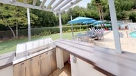 This outdoor space is thoughtfully designed for al fresco entertaining, centered around a fully equipped outdoor kitchen and a pristine swimming pool. The outdoor kitchen is the focal point, anchored beneath a clean-lined white pergola that provides partial shade while maintaining an open-air ambiance. The kitchen features a U-shaped stainless steel configuration with multiple built-in grills, generous counter space with smooth composite surfaces, and integrated cabinetry — offering everything needed for outdoor cooking and food preparation.

Surrounding the kitchen, the space opens onto a spacious patio deck that connects seamlessly to the pool area. The pool is generously sized and features a classic tiled border, with lounge chairs and vibrant blue umbrellas arranged poolside for comfortable relaxation. A balcony overlooks the pool from an adjacent structure, adding a sense of vertical dimension to the space.

The landscaping throughout is lush and well-maintained, with manicured lawns, neatly trimmed hedgerows, mature trees, and tropical palms that create a private, resort-like atmosphere. Natural light floods the entire area, enhancing the outdoor ambiance. Together, these elements compose an outdoor environment well-suited for both casual gatherings and larger-scale entertaining.