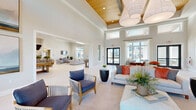 Located in Palmetto, FL, this single-floor community clubhouse offers a generous and thoughtfully designed gathering space. The architecture is defined by soaring vaulted ceilings with elegant wood-paneled tray accents, large-scale pendant lighting, and an abundance of natural light streaming through floor-to-ceiling windows and transom windows positioned high along the walls. Light-toned herringbone-patterned tile flooring flows seamlessly throughout the entire space, reinforcing the bright, airy atmosphere.

The main lounge area features multiple seating arrangements with coastal-inspired furnishings — slatted wood armchairs, plush neutral sofas, and woven area rugs — all oriented to encourage social interaction. Floor-to-ceiling glass doors open directly to an outdoor pool area, blending indoor and outdoor living.

A dedicated game room offers residents a pool table and a shuffleboard table, complemented by additional lounge seating and contemporary wall art. Adjacent to this is a well-appointed social dining and bar area, anchored by a live-edge communal table with bar-height seating, pendant lighting, and a built-in bar counter. A separate dining nook features a live-edge wood dining table with leather chairs beneath dramatic double-height windows. The overall ambiance is clean, modern, and welcoming, with a distinct Florida coastal aesthetic.