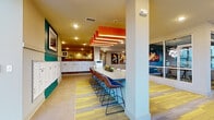 Located at 2818 Cullman Ave in Charlotte, NC, this single-floor community lobby space combines bold design with practical amenities. The entrance opens into a expansive, open-concept common area with soaring double-height ceilings and richly toned wood-look tile flooring that establishes a warm, welcoming ambiance throughout. A striking lounge area features teal velvet accent chairs, patterned curtains, and large windows that invite an abundance of natural light, complemented by a wall-mounted TV and a compact beverage station. Adjacent to the lounge, a communal bar-height table with leather saddle chairs sits beneath a dramatic cascade of illuminated orange ceiling panels, creating a vibrant co-working or social hub. A dedicated conference room showcases a bold teal accent wall, a sculptural floral chandelier, a hardwood conference table with rolling chairs, and windows overlooking the exterior — ideal for private meetings or collaborative sessions. The mail room features rows of white resident mailboxes accented by reclaimed wood paneling and teal wainscoting, adding a rustic-modern contrast. Patterned carpeting with yellow and gray tones threads through key areas, tying the color palette together cohesively. The overall design balances energetic, colorful aesthetics with functional, resident-focused spaces.